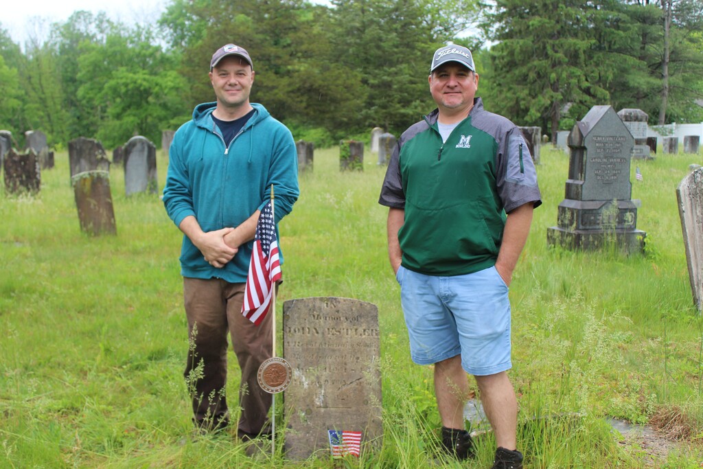 Two men stand in a grassy cemetery beside an old gravestone adorned with small American flags. Trees and more gravestones are in the background.
