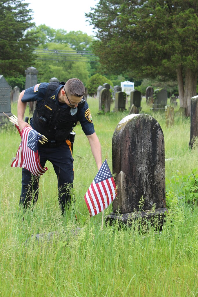 A police officer in uniform places American flags at a grave in a cemetery. The scene is solemn, with tall grass and tombstones in the background.