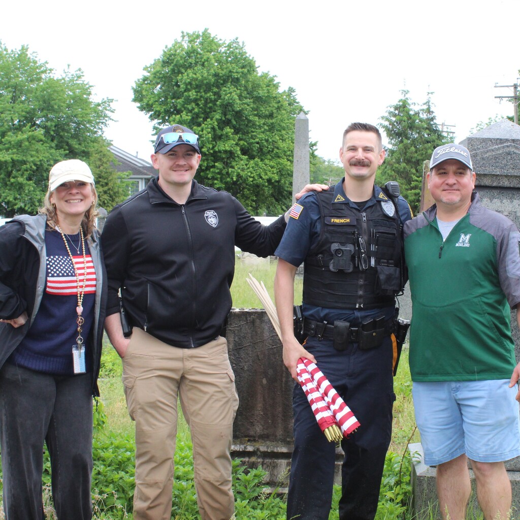 Four people stand together outdoors, smiling. One person in uniform holds rolled U.S. flags. The setting is grassy, with trees and stone monuments. Casual, friendly vibe.