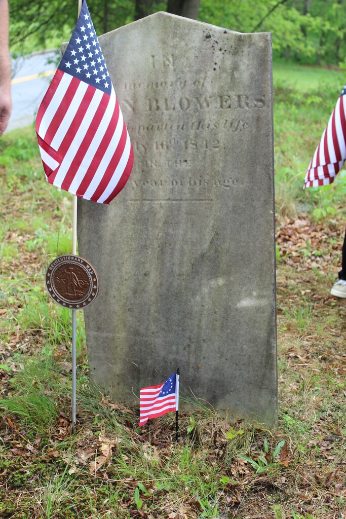 A weathered gravestone stands amidst green grass, flanked by two American flags and a Revolutionary War medallion, conveying a sense of honor.