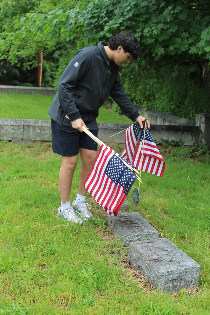 A person is placing American flags on a grave marker in a grassy cemetery, surrounded by lush greenery, conveying a solemn and respectful tone.