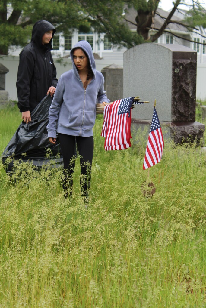 Two people in a grassy cemetery, one holding U.S. flags and the other with a trash bag. Overcast day conveying a solemn mood.