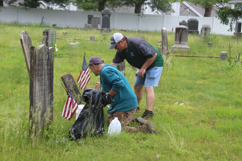 Two men clean an old cemetery, placing small American flags on tombstones. The grass is overgrown, and the scene is calm and respectful.