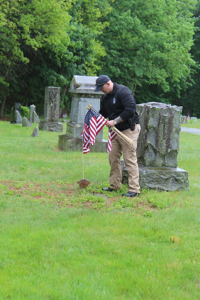A person places small American flags in front of a gravestone in a lush, green cemetery, evoking a sense of respect and remembrance.