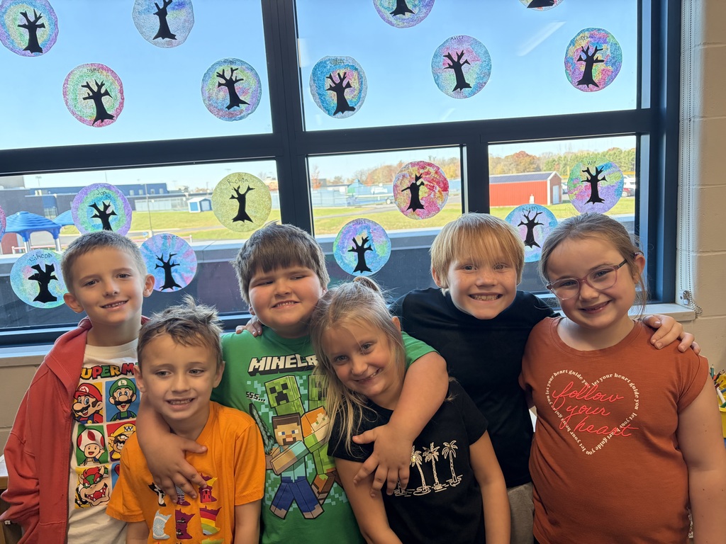 Visual Description: A bright indoor photograph of elementary-aged students standing side-by-side in a classroom, smiling with their arms wrapped around each other's shoulders in a friendly embrace.