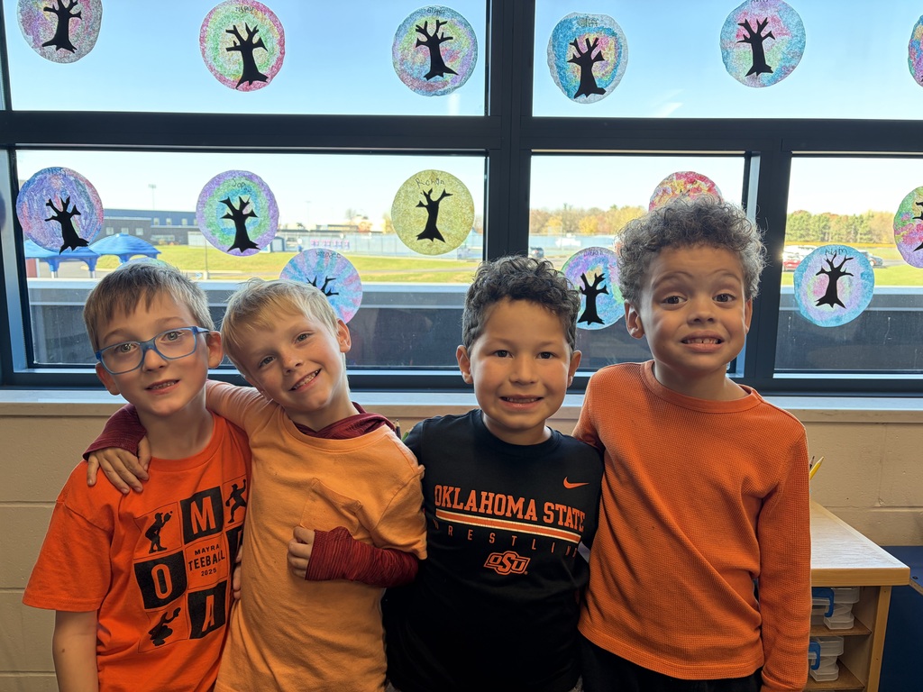 Visual Description: A bright indoor photograph of elementary-aged students standing side-by-side in a classroom, smiling with their arms wrapped around each other's shoulders in a friendly embrace.