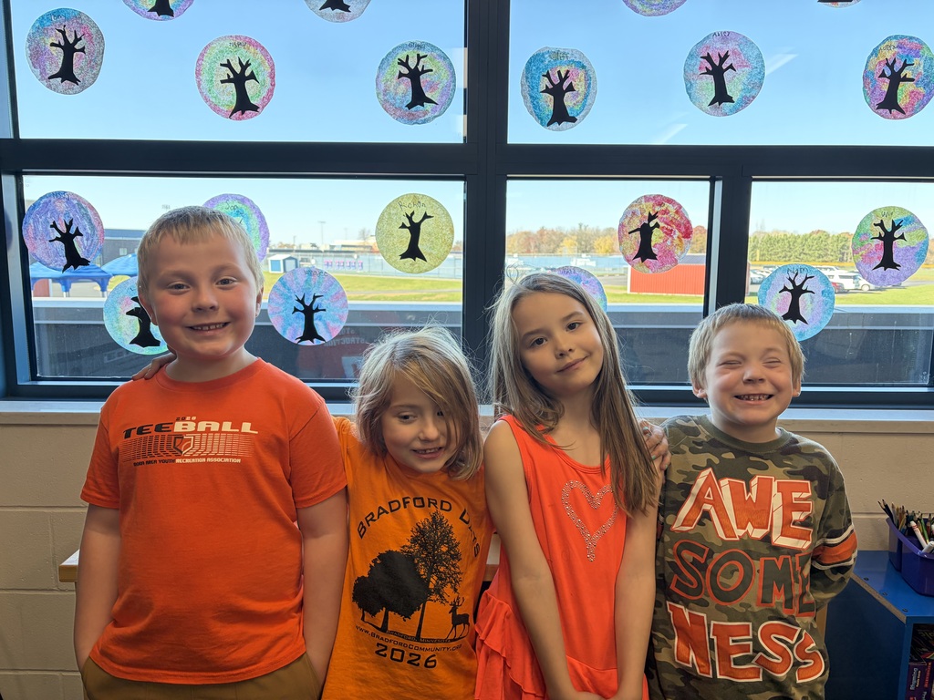 Visual Description: A bright indoor photograph of elementary-aged students standing side-by-side in a classroom, smiling with their arms wrapped around each other's shoulders in a friendly embrace.