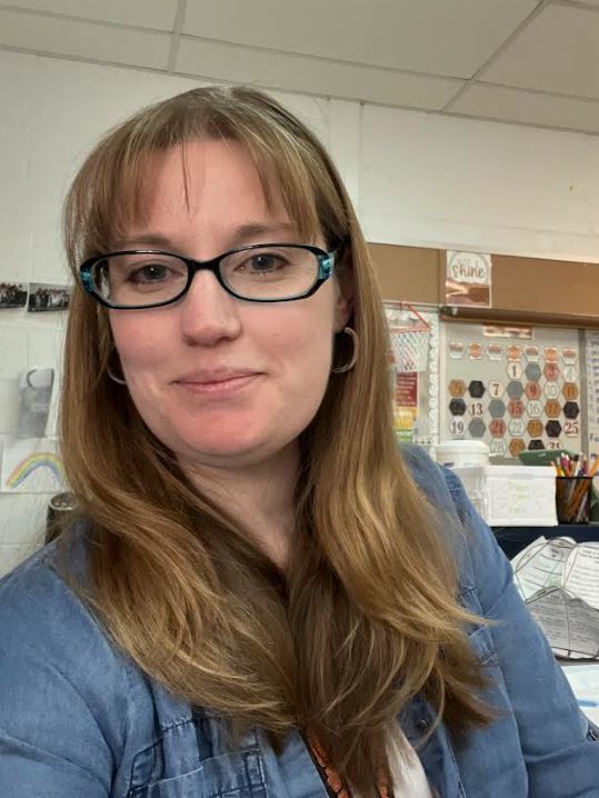Portrait of a woman with long light brown hair and glasses smiling in a classroom, with bulletin boards and educational materials visible in the background.