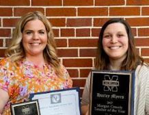 Two educators stand smiling in front of a brick wall, holding plaques and a certificate recognizing Teacher of the Year and Service Personnel of the Year achievements.