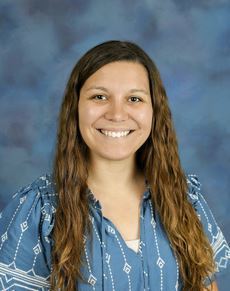 Portrait of a smiling woman with long brown hair wearing a blue patterned blouse, posed against a blue studio background.