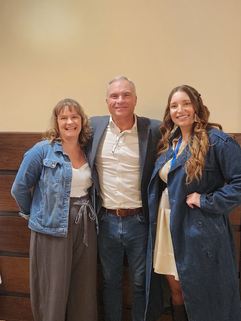 Two women and a man smiling and standing together against a tan and wood wall.