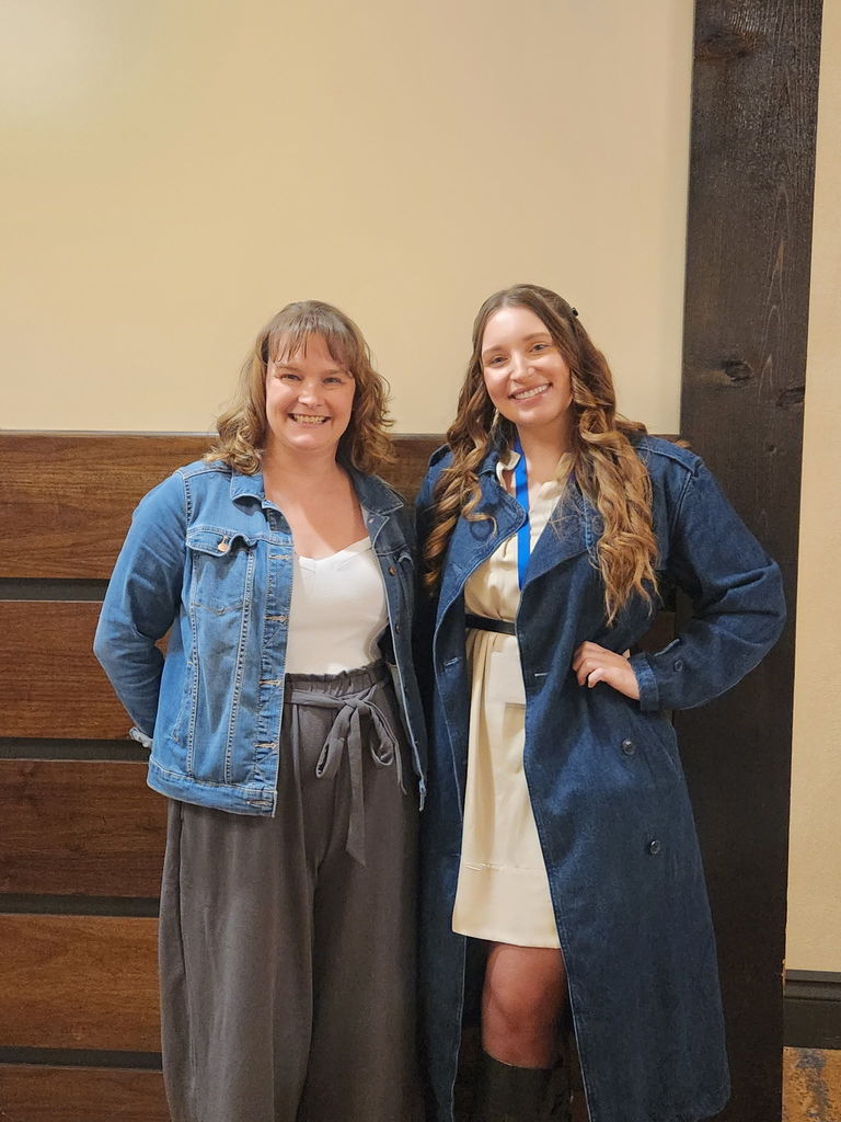 Two women smiling and standing together against a tan and wood wall.