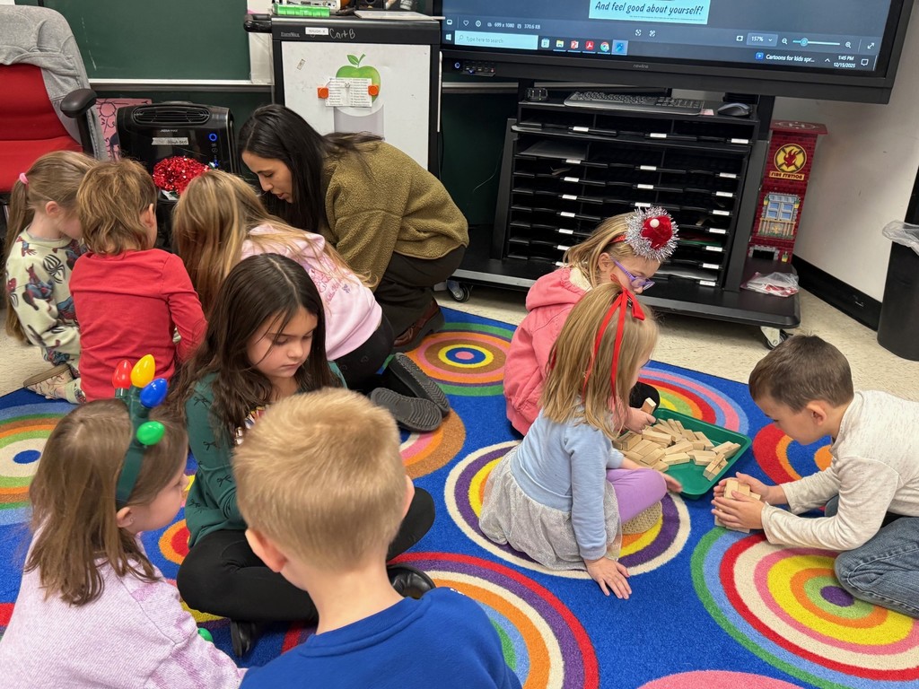 Students are sitting on the floor playing a game of Jenga. The teacher is helping. 