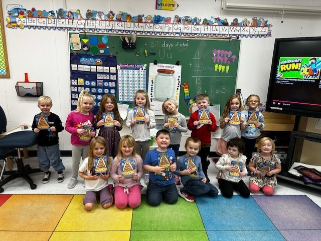 A PreK class is holding their pizza cards in front of themselves. There are 8 students standing in the back and 6 kneeling in the front.