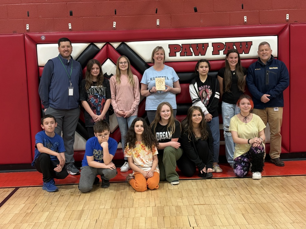 Three staff members smiling with a group of students, one staff holding an award plaque indoors.