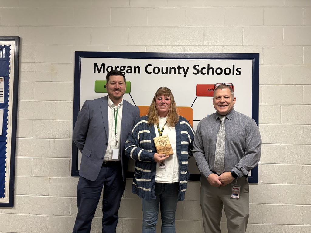 Three staff members smiling, one holding an award plaque indoors.