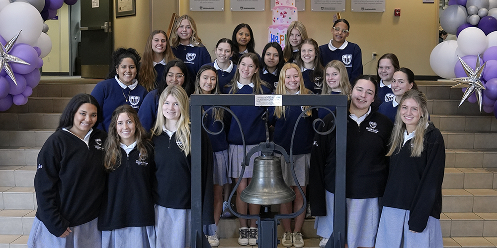 group of 21 female students standing on the steps in a foyer along with a large historic bell