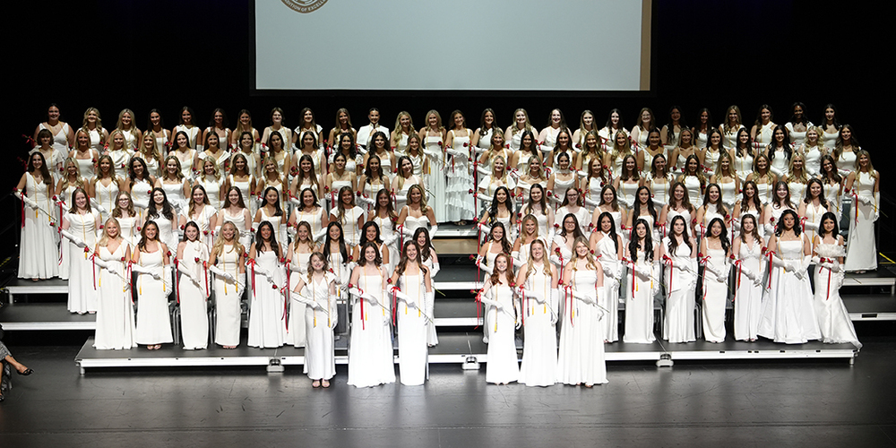 large group of graduates in long white gowns and white gloves standing on stage during their graduation ceremony