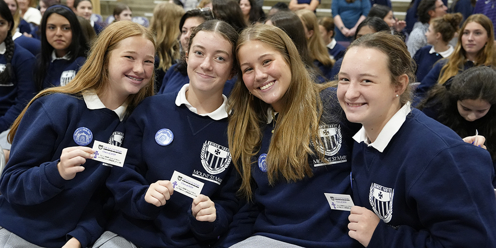 four young women in navy school sweatshirts holding up Mercy Cards and smiling at camera