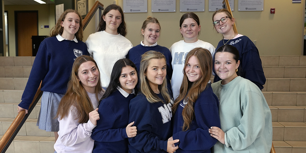 group of 10 female high school students standing on steps in school foyer and smiling at camera