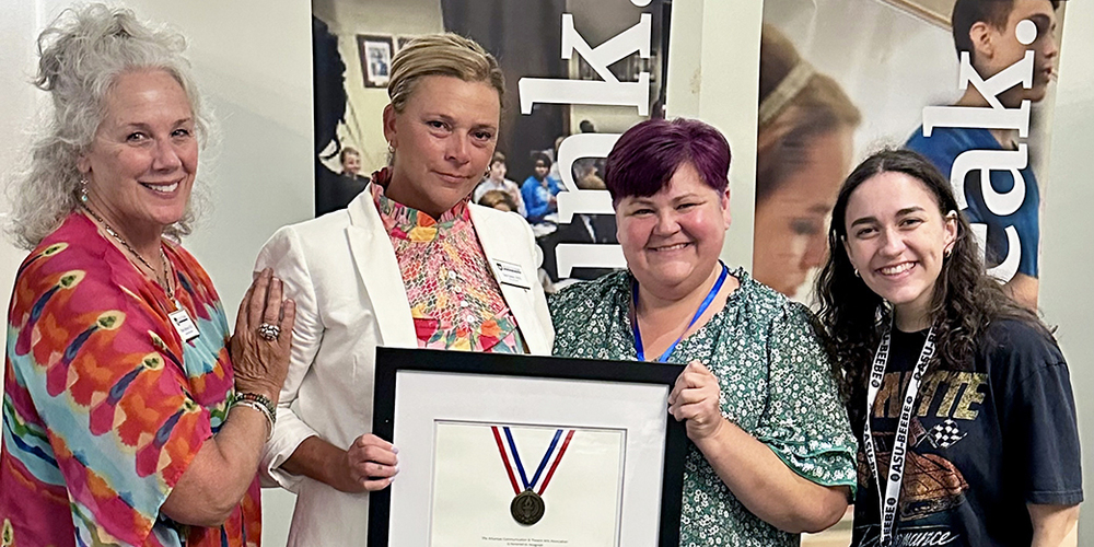 four women smiling at camera with the center two holding a framed award