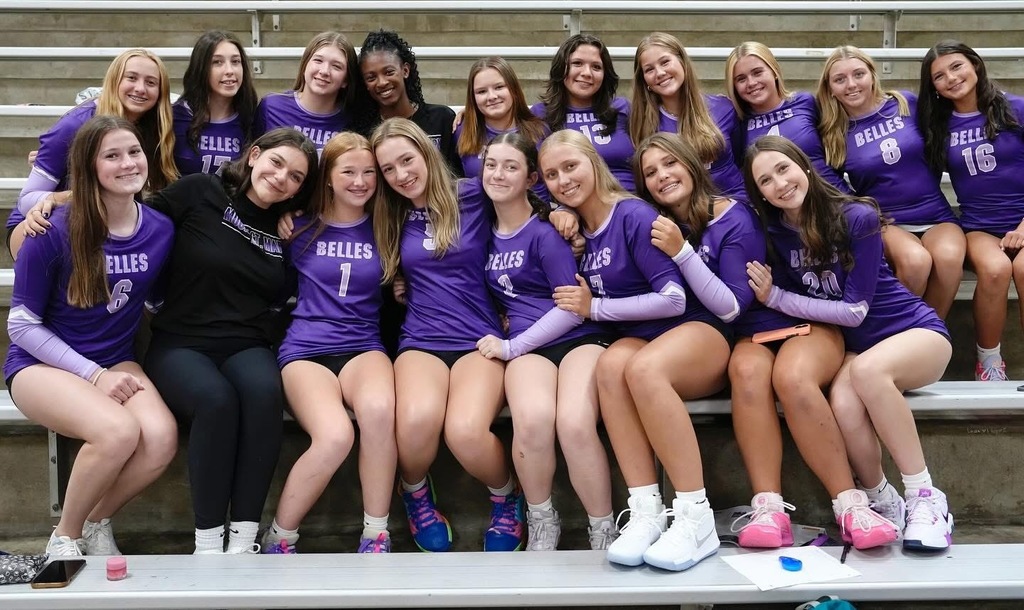 large group image of female volleyball players in purple uniforms sitting in bleachers of gymnasium