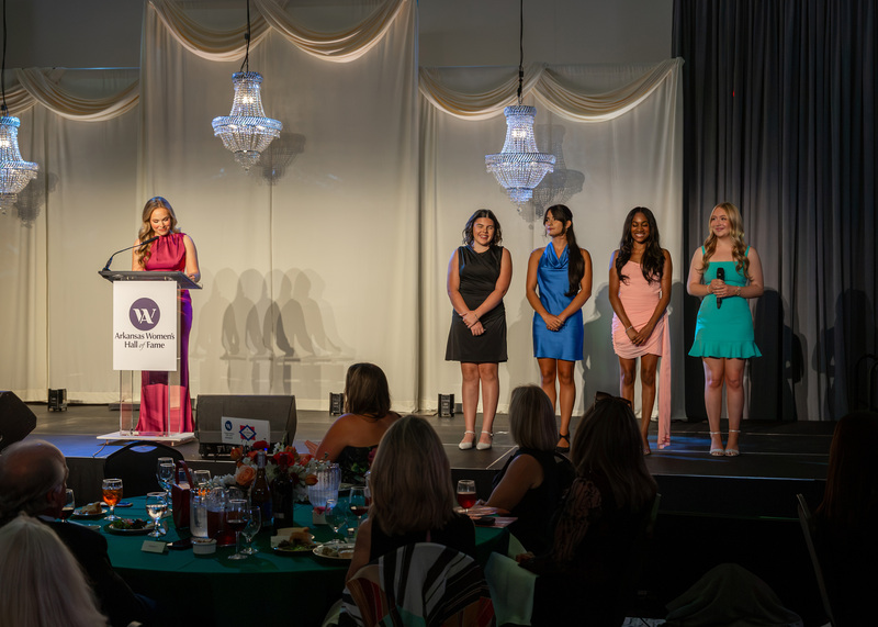 five women standing on a stage with one speaking at a podium