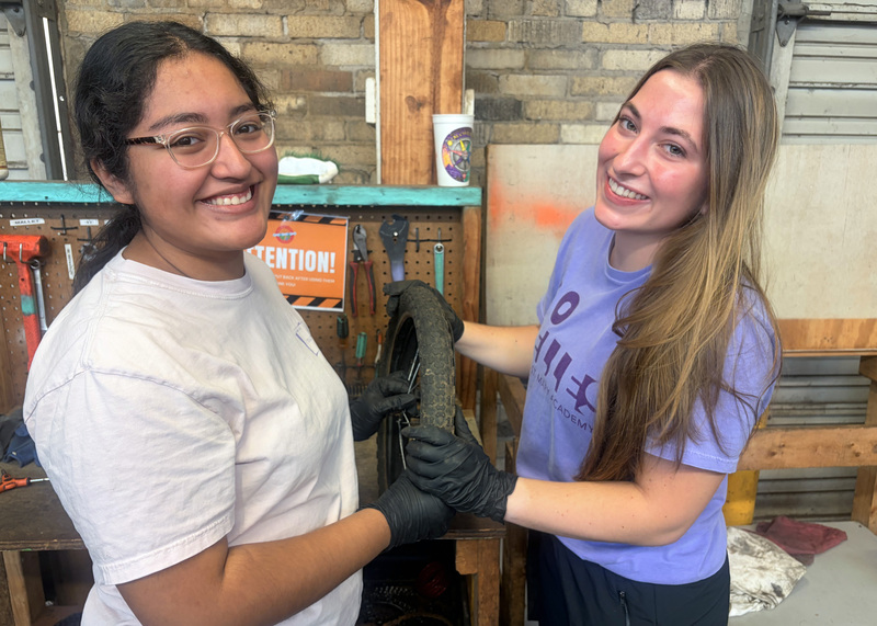 two students holding a bicycle wheel in a repair shop