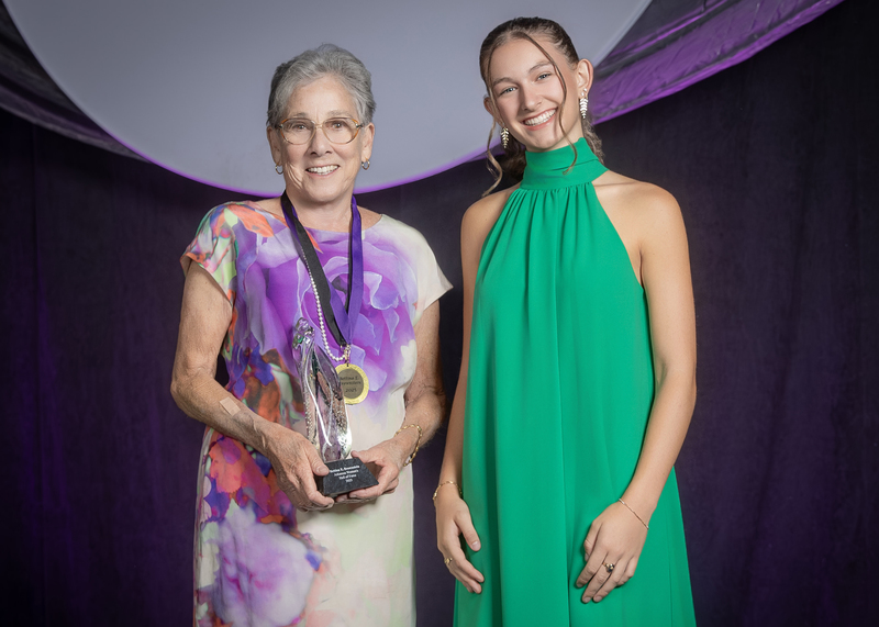 two women standing on stage, one holding an award, in front of dark purple draping