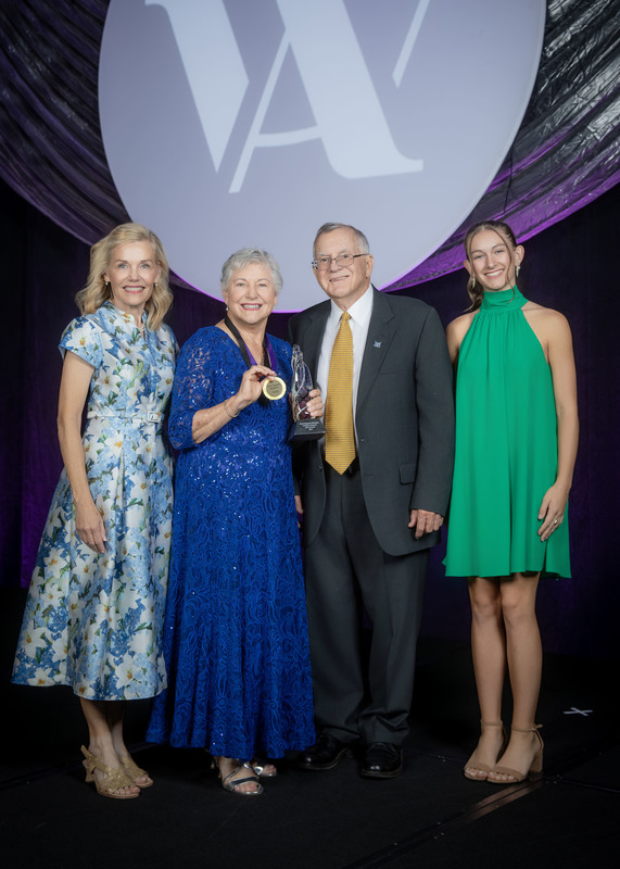 four individuals standing on stage, one holding an award, in front of dark purple draping