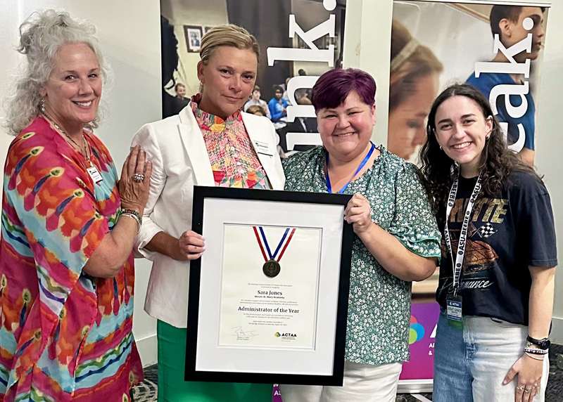 four women smiling at the camera with the center two women holding a large framed award