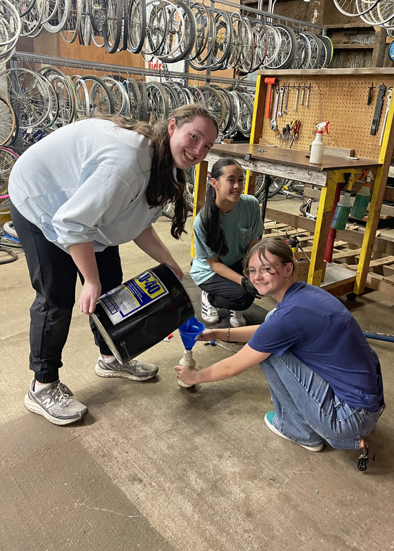 three young women in a bike shop pouring WD-40 into a smaller container using a funnel
