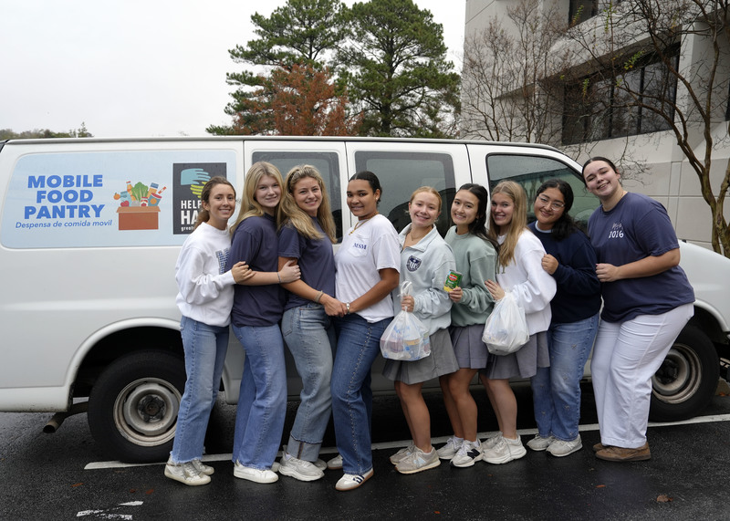 nine girls standing with plastic bags of canned goods in front of mobile food pantry van