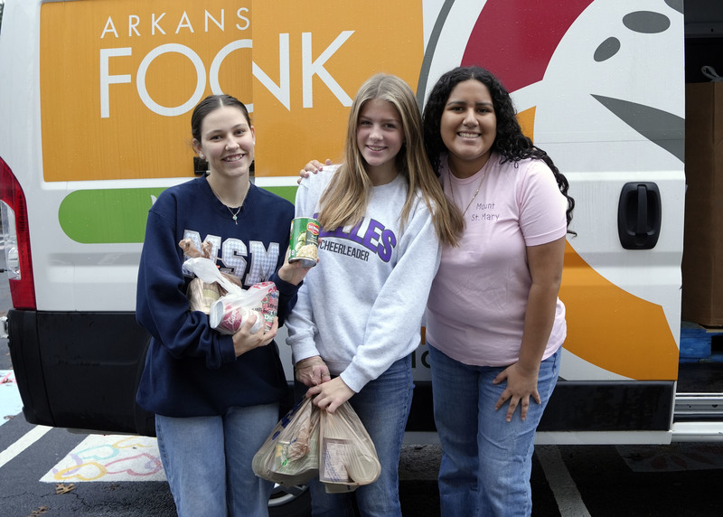 three girls standing with plastic bags of canned goods in front of Foodbank truck