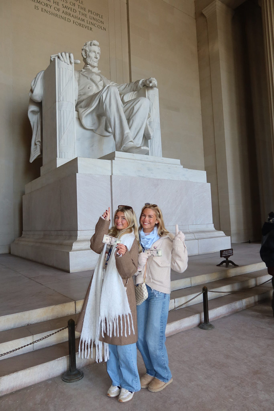 two students standing in front of the Lincoln Memorial holding a $5 bill