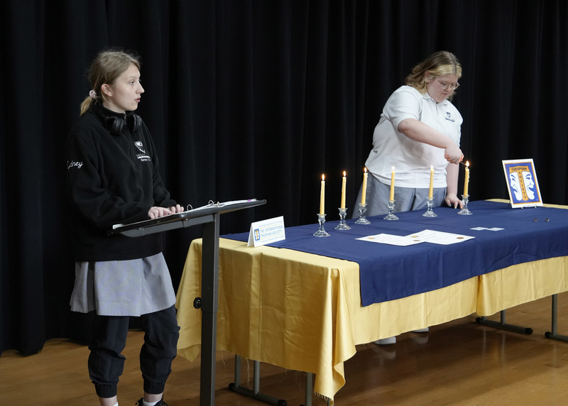 student speaking at a podium while another one lights a candle on a nearby table
