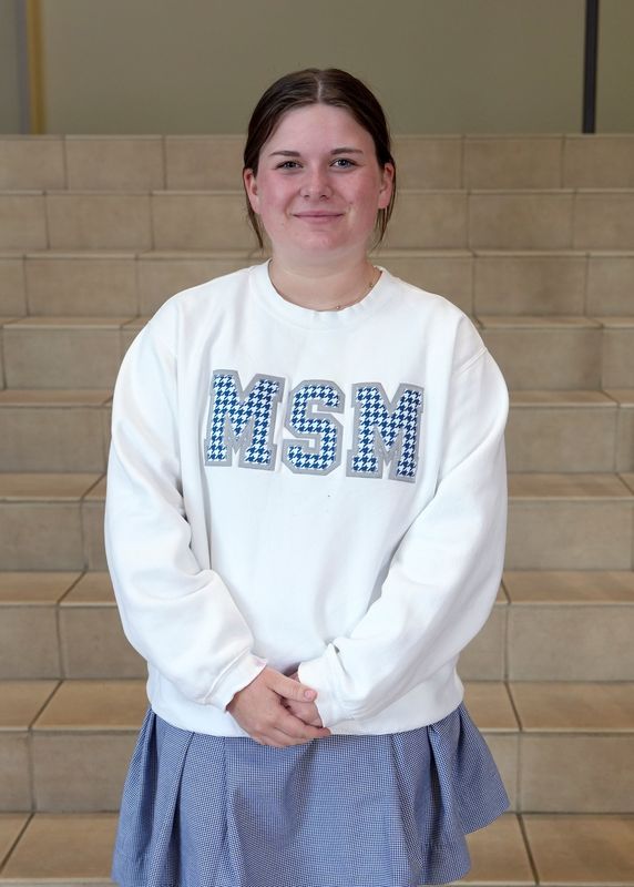 a female student standing on the steps in the school's foyer smiling at the camera