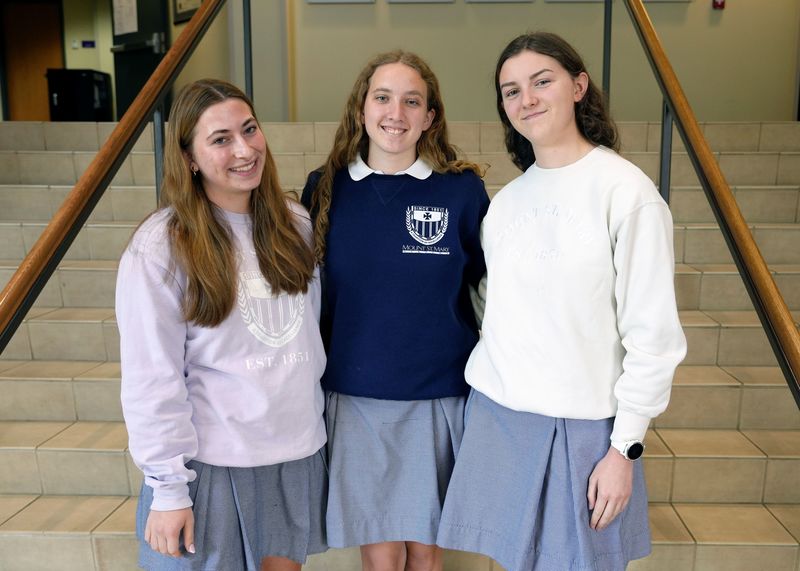 three female students standing on the steps in the school's foyer smiling at the camera