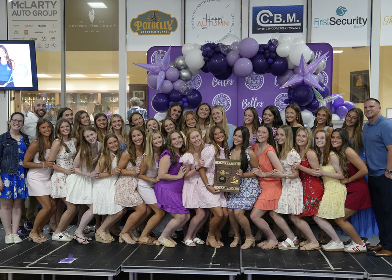 large group of students and their two coaches standing on a stage and smiling at the camera with the championship plaque