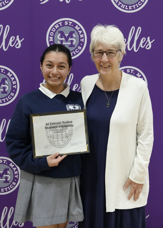 a student and an adult standing in front of a purple backdrop, holding an award certificate, and smiling at the camera