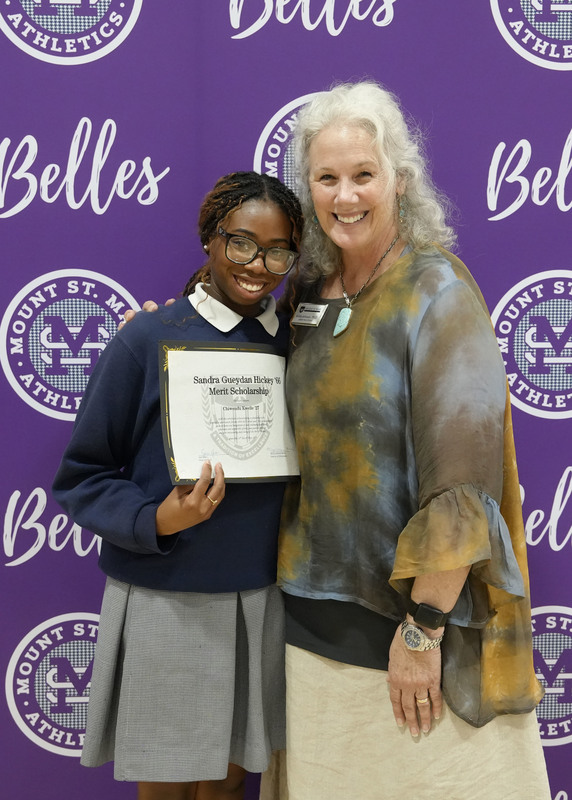 a student and an adult standing in front of a purple backdrop, holding an award certificate, and smiling at the camera