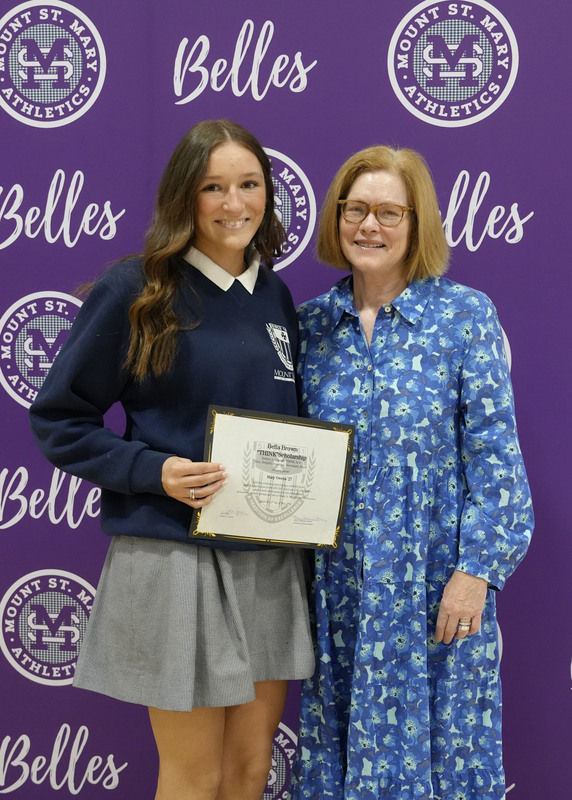 a student and an adult standing in front of a purple backdrop, holding an award certificate, and smiling at the camera
