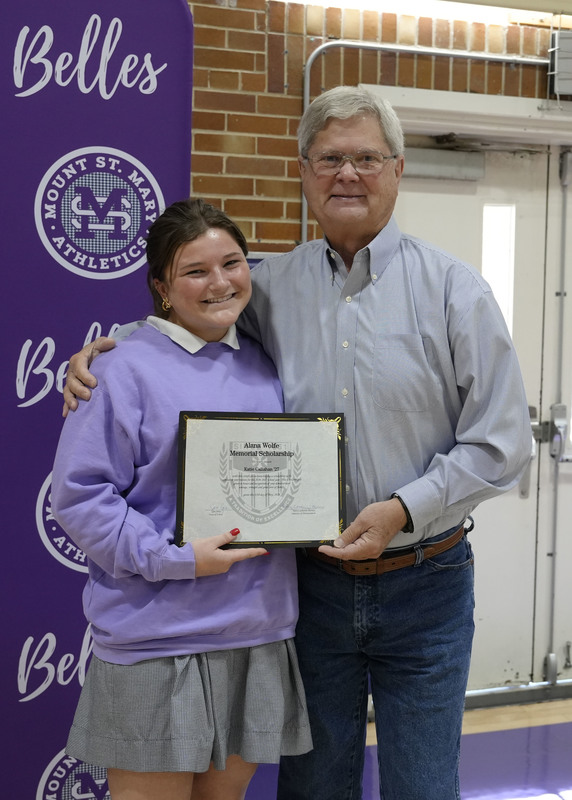 a student and an adult standing in front of a purple backdrop, holding an award certificate, and smiling at the camera