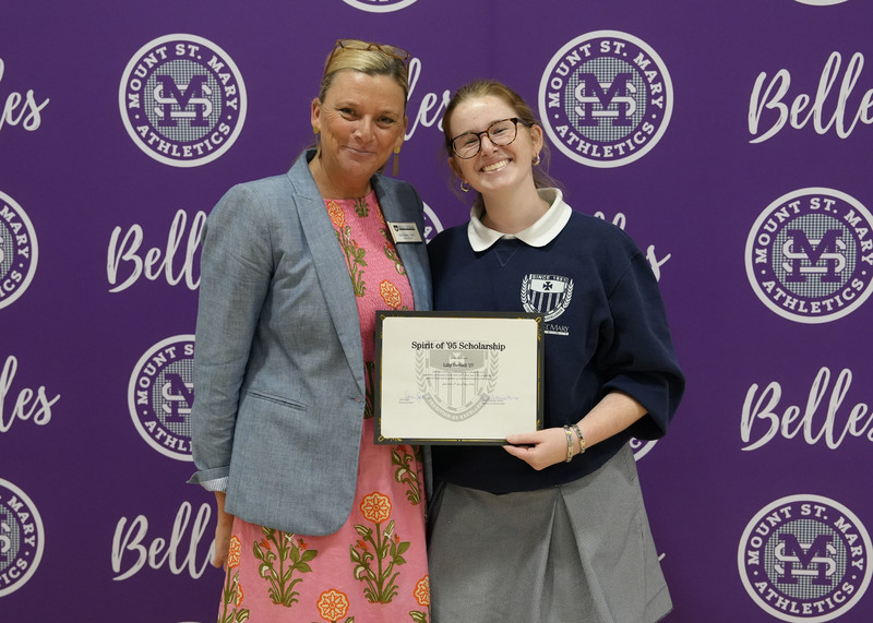 a student and an adult standing in front of a purple backdrop, holding an award certificate, and smiling at the camera