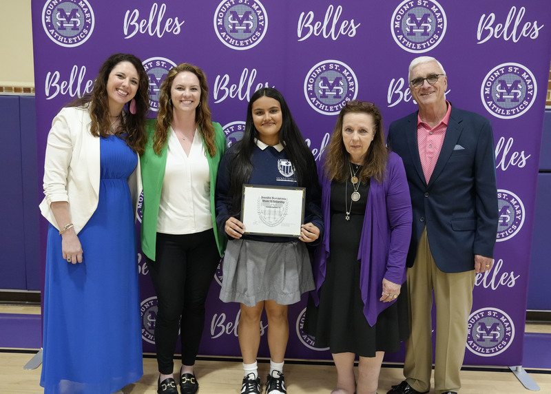 a student with four adults standing in front of a purple backdrop, holding an award certificate, and smiling at the camera