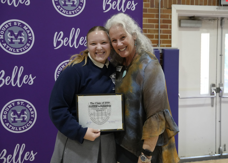 a student and an adult standing in front of a purple backdrop, holding an award certificate, and smiling at the camera