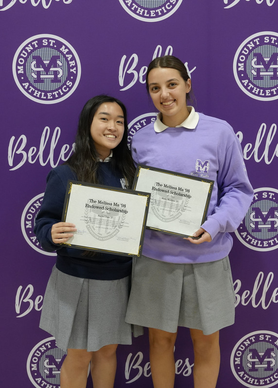 two students standing in front of a purple backdrop, holding award certificate,s and smiling at the camera