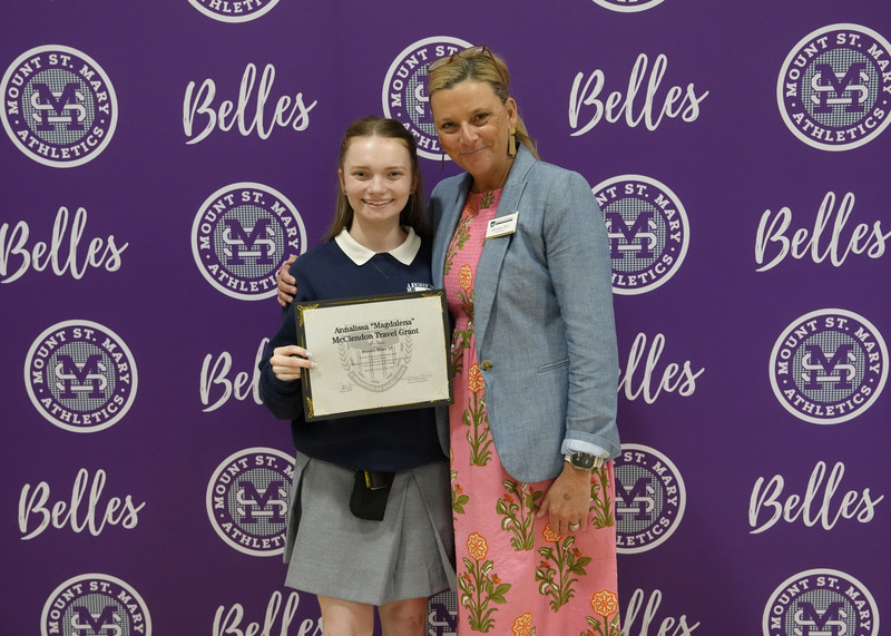 a student and an adult standing in front of a purple backdrop, holding an award certificate, and smiling at the camera