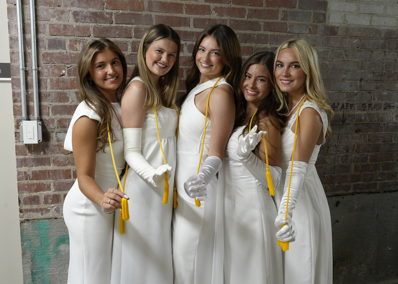 five young women in long white gowns and white gloves standing in front of a brick wall and smiling for the camera