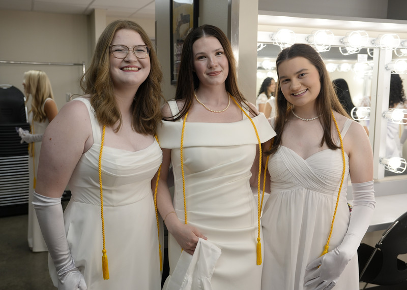 three young women in long white gowns and white gloves standing in a dressing room and smiling for the camera